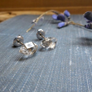 May include: A pair of clear, faceted crystal stud earrings with silver posts. The earrings are diamond-shaped and reflect light. The background is a blue textured surface with a sprig of lavender.