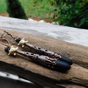 May include: A pair of long, black and copper earrings with a white bone-like charm. The earrings feature a detailed, intricate design with a black, rounded tip.