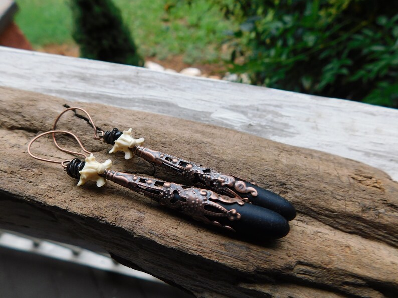 May include: A pair of long, black and copper earrings with a white bone-like charm. The earrings feature a detailed, intricate design with a black, rounded tip.
