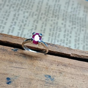 Op de afbeelding: Een zilveren ring met een grote, ovaal geslepen robijn. De robijn heeft een dieprode kleur en is gezet in een vier-klauwzetting. De ring wordt gepresenteerd op een houten oppervlak.