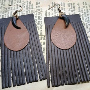 May include: A pair of earrings featuring dark brown leather fringe attached to a tan leather teardrop shape. Each earring is accented with a curved, dark gray claw-like element at the top, suspended from a bronze-toned metal hook wire.