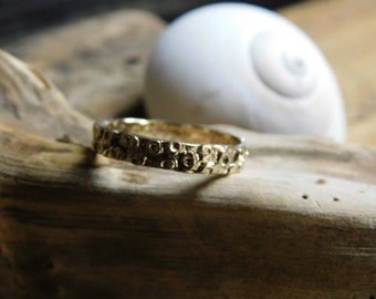May include: A textured gold ring with a repeating pattern of small circles. The ring is set on a piece of driftwood, with a white seashell in the background. The ring appears to be handmade.