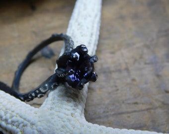 May include: A black ring with a dark purple gemstone set in a claw setting. The ring is made of a textured metal and has a unique design.