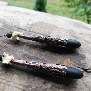 May include: A pair of long, black and copper earrings with a white bone-like bead at the top. The earrings have a delicate, intricate design.