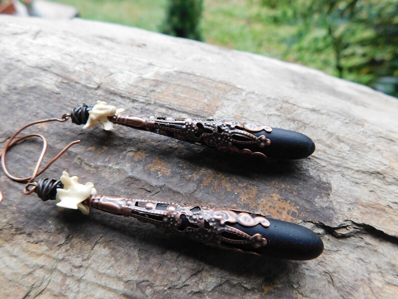 May include: A pair of long, black and copper earrings with a white bone-like bead at the top. The earrings have a delicate, intricate design.