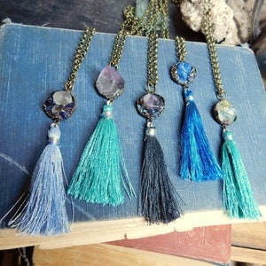 May include: Five necklaces with different colored gemstone pendants and tassel charms. The tassels are blue, teal, black, and turquoise.