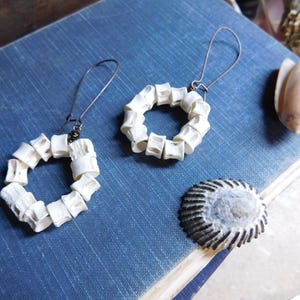 May include: A pair of white bone earrings with a circular shape. The earrings are made of small, white bone pieces that are connected together to form a circle. The earrings are hanging from silver hooks.