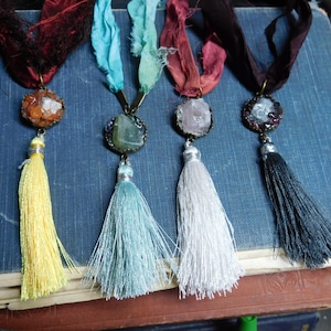 May include: Four necklaces with different coloured tassels and crystal pendants. The crystals are set in bronze-coloured metal settings. The tassels are yellow, blue, white, and black.