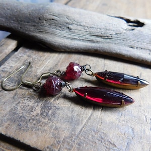 May include: A pair of dangle earrings with a dark red glass teardrop shape and a smaller dark red faceted bead. The earrings have a gold tone metal setting and a hook closure.