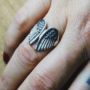 May include: A silver ring with a detailed design of two angel wings.