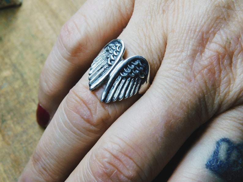 May include: A silver ring with a detailed design of two angel wings.