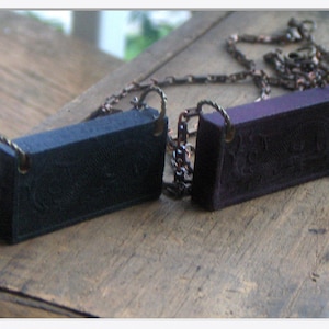 May include: Two rectangular pendants with a chain. The pendants are made of dark blue and purple resin and have a textured surface. The chain is made of copper and has a vintage look.