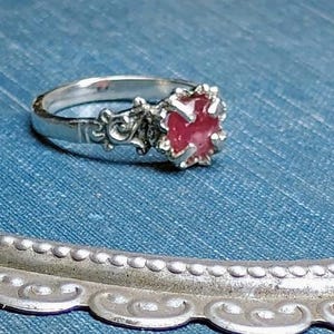 May include: A silver ring with an ornate band and a central, red gemstone. The gemstone is set in a decorative, pronged setting. The ring is displayed on a blue denim surface.