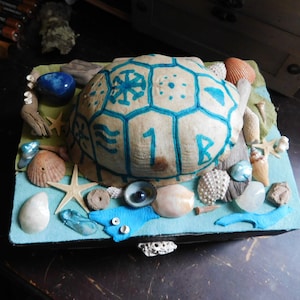 Könnte beinhalten: Eine dekorative Box mit einem bemalten Schildkrötenschalen-Deckel. Die Box ist gefüllt mit Muscheln, Seesternen und anderen Meeres-Dekorationen. Der Deckel ist in Blau und Weiß mit einem Schneeflocken-Muster bemalt und die Zahl "18" ist darauf geschrieben.