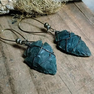 May include: A pair of earrings featuring dark green, arrowhead-shaped stone pendants wrapped in thin, dark metal wire. The pendants are attached to bronze-toned metal ear wires, each accented with small, round metallic beads near the top hook.