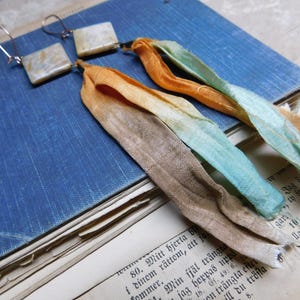 May include: Earrings with square, off-white pendants and long, colorful silk ribbons in shades of brown, orange, and blue. The earrings rest on a blue book and vintage paper.