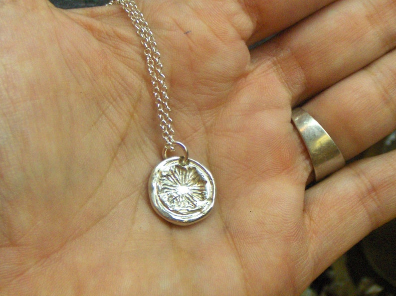 May include: A silver necklace with a round pendant featuring a detailed floral design. The pendant is slightly raised and has a textured surface.