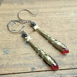 May include: A pair of gold-toned earrings with red glass teardrop accents. The earrings feature intricate details and black beads.