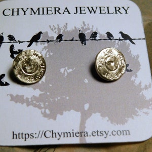 以下が含まれることがあります： 金色のスタッドピアス、円形デザイン。ピアスは「CHYMIERA JEWELRY」の文字とワイヤー上の鳥のシルエットが描かれた白いカードに展示されています。ウェブサイトのアドレスhttps://chymiera.etsy.comも表示されています。