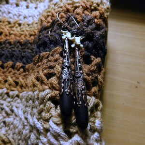 May include: A pair of black and copper earrings with a bone-like design. The earrings are hanging from a crocheted fabric background.
