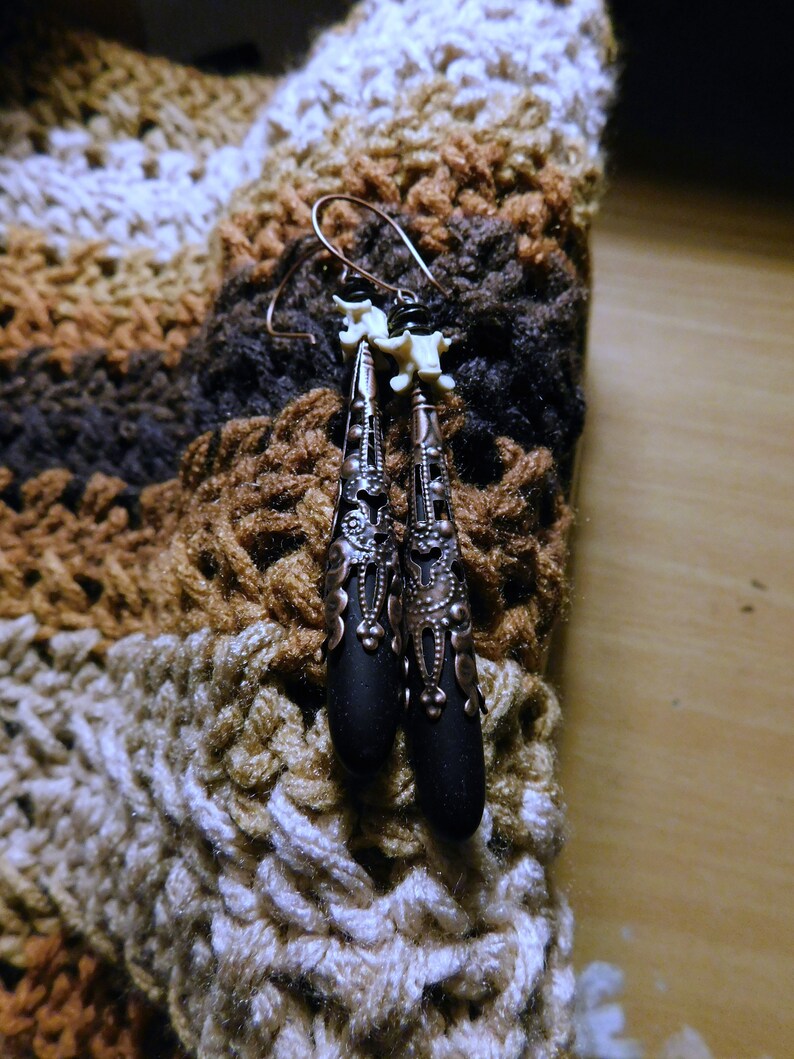 May include: A pair of black and copper earrings with a bone-like design. The earrings are hanging from a crocheted fabric background.