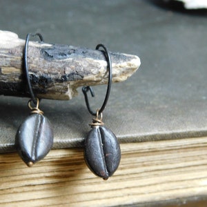 May include: A pair of black hoop earrings with brown coffee bean charms hanging from them. The earrings are displayed on a wooden surface.