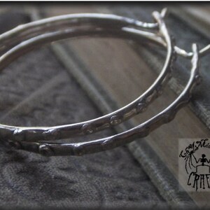 May include: Two silver hoop earrings with a textured, twig-like design. The earrings are displayed on a textured gray surface. The earrings have a unique, organic aesthetic, perfect for adding a touch of nature-inspired style to any outfit.