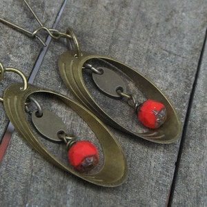 May include: A pair of brass earrings with a unique design. Each earring features a circular shape with a smaller circle inside, and a red bead hanging from the bottom. The earrings are a statement piece and would be a great addition to any jewelry collection.