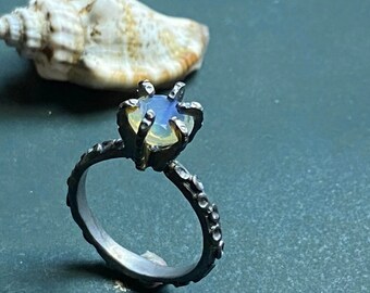 May include: A silver ring with a textured band and a prong setting holding a light blue and yellow gemstone. The ring is set against a dark green background with a seashell.