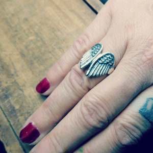 May include: A silver ring with a detailed wing design. The ring is worn on a finger with red nail polish.