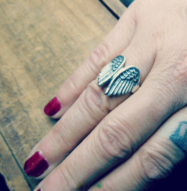 May include: A silver ring with a detailed wing design. The ring is worn on a finger with red nail polish.
