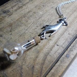 May include: A silver hand charm necklace with a small glass bottle filled with seashells. The bottle is attached to the hand charm with a cork.