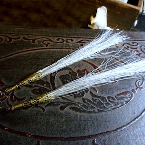 May include: A pair of handmade earrings featuring long, white, feathery strands. The earrings have gold-colored filigree caps and ear wires. The earrings are displayed on a dark, textured surface with an embossed design.