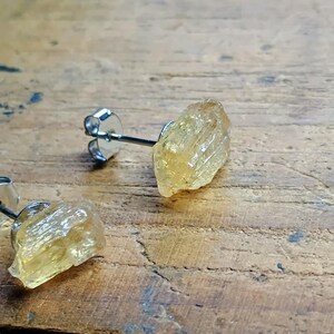 Puede incluir: Un par de pendientes de bot&oacute;n con piedras preciosas de citrino amarillo dorado en bruto. Los pendientes tienen postes plateados y cierres de mariposa. Las piedras preciosas tienen una textura rugosa y natural, sobre un fondo de madera.