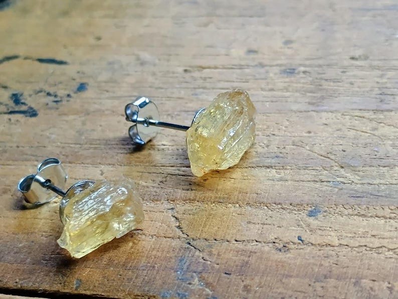 Puede incluir: Un par de pendientes de bot&oacute;n con piedras preciosas de citrino amarillo dorado en bruto. Los pendientes tienen postes plateados y cierres de mariposa. Las piedras preciosas tienen una textura rugosa y natural, sobre un fondo de madera.