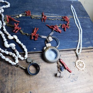 May include: Three necklaces with different charms. One necklace is made of white seashells, another is made of red coral, and the third is made of white pearls with a gold ship's wheel pendant.