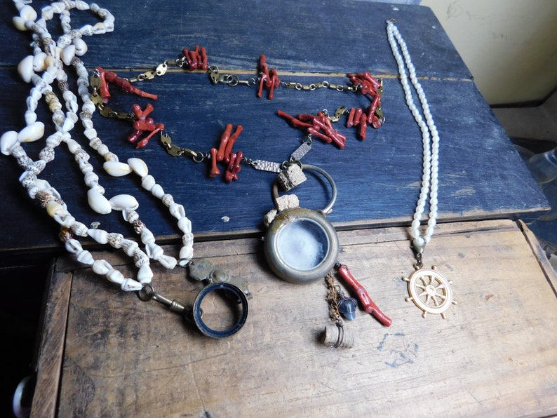 May include: Three necklaces with different charms. One necklace is made of white seashells, another is made of red coral, and the third is made of white pearls with a gold ship's wheel pendant.