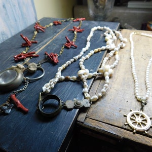 May include: Three necklaces on a wooden surface. One necklace is made of red coral beads, another is made of white shell beads, and the third is made of white pearls with a gold ship's wheel pendant.