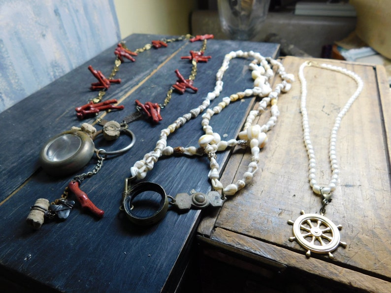 May include: Three necklaces on a wooden surface. One necklace is made of red coral beads, another is made of white shell beads, and the third is made of white pearls with a gold ship's wheel pendant.