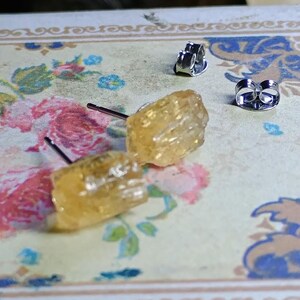 Puede incluir: Un par de pendientes de bot&oacute;n de citrino en bruto con postes y cierres de plata. Los pendientes tienen un aspecto rugoso y texturizado y un color amarillo dorado c&aacute;lido. Los pendientes se muestran sobre una superficie con estampado floral.