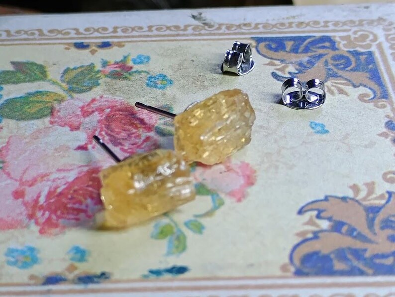 Puede incluir: Un par de pendientes de bot&oacute;n de citrino en bruto con postes y cierres de plata. Los pendientes tienen un aspecto rugoso y texturizado y un color amarillo dorado c&aacute;lido. Los pendientes se muestran sobre una superficie con estampado floral.