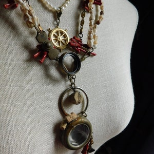 May include: A layered necklace with a variety of charms, including a ship's wheel, a magnifying glass, and a small round container. The necklace is made of white pearls, red coral, and gold-toned metal.