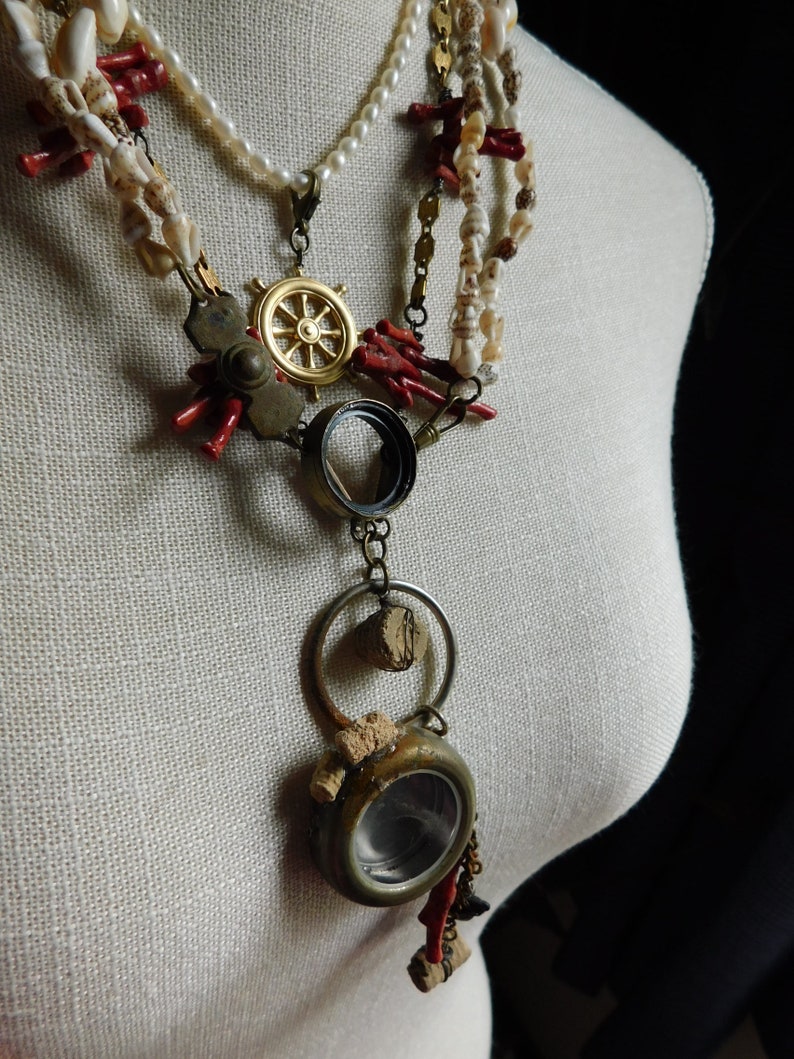 May include: A layered necklace with a variety of charms, including a ship's wheel, a magnifying glass, and a small round container. The necklace is made of white pearls, red coral, and gold-toned metal.