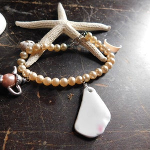 May include: A pearl necklace with a teapot charm, a white porcelain charm, and a starfish. The necklace is on a brown wooden surface.