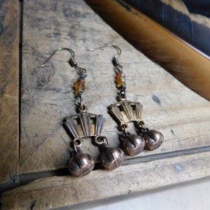 May include: A pair of gold tone earrings with a geometric design and small bells hanging from the bottom. The earrings are made with a chain and beads.