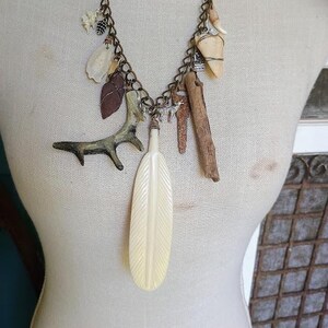 May include: A charm necklace featuring a bronze-toned chain adorned with various natural elements including a large cream-colored feather pendant, a small antler piece, a piece of driftwood, a dark stone arrowhead, and a small silver wolf charm.