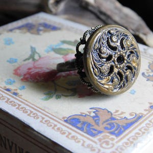May include: A vintage-style ring with an ornate, gold-toned button as its centerpiece. The button features intricate floral and swirl patterns, with a dark center stone. The ring's band has a dark, antique finish, and the ring is set on a decorative box.