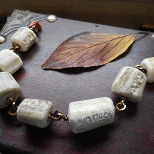 May include: A white antler bracelet with the words "You", "Are", "Lost", "Wander", and "Old" carved into the beads. The bracelet is on a wooden surface with a brown leaf.