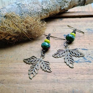 May include: A pair of handcrafted earrings featuring bronze-colored leaf charms and green and yellow beaded accents. The earrings have a rustic, nature-inspired design, with a hook closure. The earrings are displayed on a wooden surface.