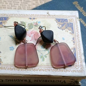 May include: A pair of handmade earrings featuring black stone beads and translucent, rectangular pink-purple glass beads. The earrings are suspended from copper wire hooks and are displayed on a vintage book.